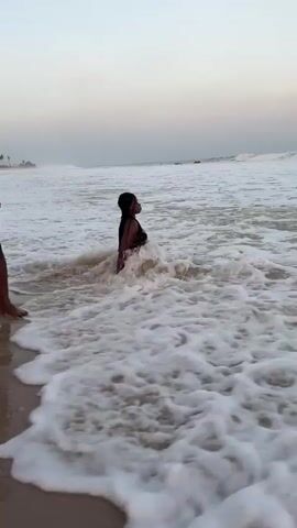 A woman in a bikini sits in foamy ocean water, smiling. A person in patterned shorts stands in the foreground, slightly to the left. The ocean stretches to the horizon, with waves breaking in the distance. The sky is overcast, with hints of pale colors.
