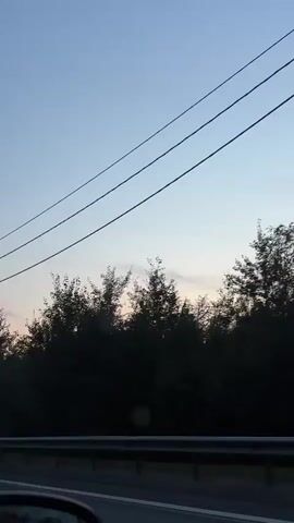 This is a vertical, first-person video taken from a vehicle driving along a road. The time of day appears to be dusk or dawn, with a clear sky transitioning from light blue to a soft pinkish hue near the horizon. A dense line of dark trees forms the background. Two parallel power lines stretch diagonally across the upper portion of the frame, disappearing off-screen to the right. In the lower foreground, a portion of the vehicle's interior is visible, including a side mirror reflecting the scenery. A metal guardrail runs along the side of the road. The overall impression is one of a quiet, scenic drive through a wooded area during twilight.