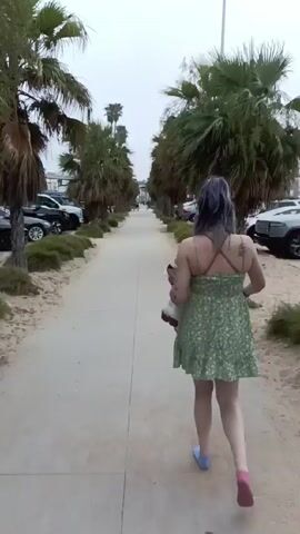 A woman walks away from the camera on a concrete path toward the ocean. The path is lined with palm trees on both sides. Sand and parked cars can be seen adjacent to the path. She is wearing a short, light-green, floral dress with thin straps. She has blue hair, and tattoos on her back and arms. She is carrying a white tote bag and what looks like a clear plastic cup. She is wearing blue socks, and appears to be barefoot. The weather looks overcast.