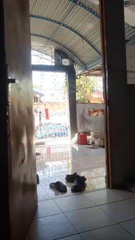 A stationary, eye-level shot shows an interior tiled floor looking out through an open wooden doorway. Several pairs of dark flip-flops or sandals are scattered on the light-colored tiled floor near the threshold. Beyond the doorway, a covered outdoor area with a corrugated metal roof is visible, also featuring a tiled floor. A red jerrycan and a broom are present in this outer area. Further in the background, a metal gate or fence is seen, with other structures and some foliage beyond it. The scene is brightly illuminated by daylight. No individuals are present.