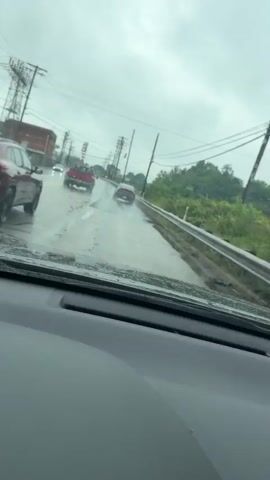 The video captures a point-of-view shot from inside a vehicle driving on a wet road under a cloudy sky. The road appears to be in a semi-urban or suburban area, with power lines and utility poles lining the right side of the road. Several vehicles are visible ahead, traveling in the same direction. The closest vehicle in front of the camera is a red SUV. Further ahead, a red pickup truck is visible, and beyond that, a dark-colored sedan. The road surface is visibly wet, with puddles and reflections indicating recent or ongoing rain. The overall atmosphere is overcast and damp, suggesting inclement weather.