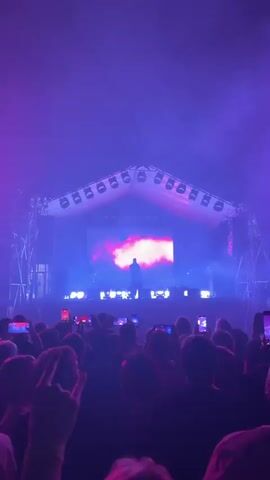A concert crowd is shown from behind, facing a stage illuminated by purple and pink lights. A performer stands center stage with a large screen behind them displaying a swirling cloud-like image. In the next scene, the stage is overwhelmed with bright yellow spotlights. The final scene shows the stage under red lighting, with the band members silhouetted in front of a screen displaying a pink abstract image. The crowd holds up numerous smartphones, recording the performance.