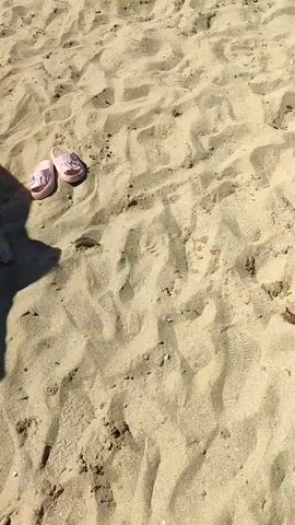 The video shows a sandy beach. In the foreground, there are two small pink sandals. The camera pans slightly to the right, revealing more of the sand and some footprints. Further back, another pair of blue sandals are visible, along with parts of beach chairs and a blue umbrella. The scene is outdoors under natural light, indicating a sunny day. There are no people clearly visible in the frame, though shadows suggest their presence.