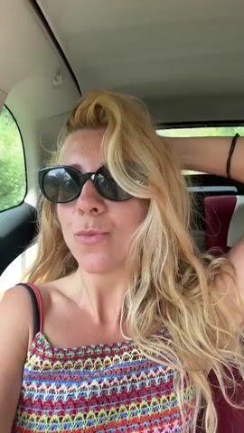 The video begins with a close-up of a woman with blonde hair and sunglasses, wearing a colorful crocheted top, sitting in the passenger seat of a car. She smiles and adjusts her hair. The scene then shifts to a blurry shot of a man in the driver's seat, also wearing sunglasses. Finally, the video shows a view from inside the car looking out through the windshield at a road surrounded by trees and greenery. The lighting suggests it is daytime, possibly late morning or early afternoon.