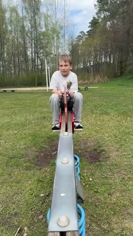 A young boy is sitting on a seesaw in a park. He is wearing a white t-shirt, gray pants, and black sneakers. The seesaw is made of metal and wood, and has blue springs underneath. The park has green grass, trees, and a sandbox in the background. The sky is cloudy.