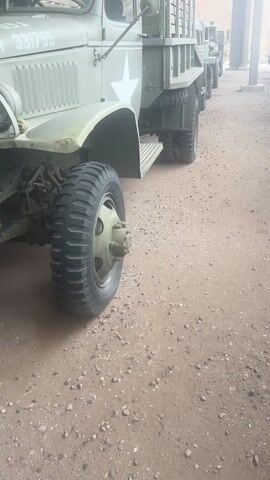 The video shows a row of vintage olive drab military trucks parked in a covered, open-sided structure with a dirt floor. The trucks have white stars painted on their sides. The first truck in the row has the number "331755" printed on the hood. The trucks appear to be in good condition. The background reveals a desert landscape with a low mountain range.  The camera moves slowly, panning from the front of the first truck toward the end of the row.