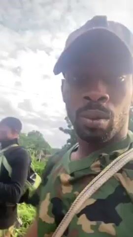 The video features three men walking in what appears to be a rural area. The man closest to the camera is wearing a camouflage shirt and cap, with a strap across his chest. He is looking directly at the camera. Behind him, two other men are walking. One is wearing a black shirt, camouflage pants, and a vest, and is carrying a rifle. The third man is wearing a yellow shirt. The background includes greenery, houses with straw roofs, and a cloudy sky. The video is shot in a handheld style, suggesting the person filming is walking along with the group. The background music is audible.