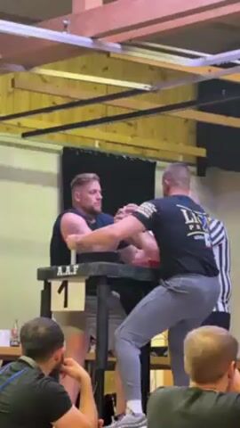 Two muscular men are engaged in an arm-wrestling match at a table marked "1". One man is wearing a black tank top and grey sweatpants, while the other is wearing a black t-shirt with "Lion Pride" written on it and grey sweatpants. A referee in a black and white striped shirt stands behind them, overseeing the match. A small crowd watches the competition. The setting appears to be a gymnasium or large hall with exposed wooden beams on the ceiling. The man in the black tank top wins.