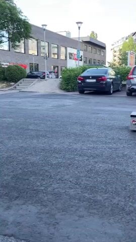 This video captures a young man skateboarding in what appears to be a public outdoor space in Karlstad, Sweden. The scene is set during daylight, with a bright, overcast sky suggesting it could be late afternoon or early evening. The foreground is dominated by a rough asphalt surface, with a low, graffiti-covered ramp or ledge placed centrally.

The main subject is the skateboarder, dressed in a light-colored short-sleeved shirt, dark shorts, and white sneakers. He is actively engaged in skateboarding, demonstrating a trick where he approaches a ledge, ollies onto it, and then rides off. His movements suggest skill and practice.

In the background, several cars are parked, including a dark sedan and a silver Volvo. Beyond the parking area, there are multi-story residential buildings with modern architecture, indicative of an urban or suburban environment. Lush green trees and bushes add to the natural scenery, contrasting with the urban elements.

There are a few other people visible in the mid-ground, walking away from the camera, seemingly unaware or unconcerned with the skateboarding activity. One couple is walking together, while another individual walks further ahead.

Visible text includes "PLAZA" spray-painted multiple times on the skateboard ledge. On the building in the background, a sign with "WILS" is visible, and another sign further down the street reads "BYGG". A street sign with a green background and white text is also present, but illegible. A scooter is parked near the buildings on the right side of the frame. The overall mood is casual and recreational, capturing a moment of urban sport.