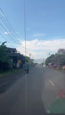 A vertical, first-person perspective video shows a road with a bright blue sky and scattered clouds above. The road stretches forward, with a white line dividing it and a yellow line on the right side. On the left side of the road, there are buildings, including a structure with a metal roof and a signpost indicating a left turn. A person on a motorcycle, wearing a blue garment, is traveling away from the camera on the left side of the road. Power lines crisscross the sky. On the right side of the road, there are more buildings, some appearing to be shops, with a sign for "AQUA" visible. The scene appears to be a semi-urban or rural area. The camera is positioned from inside a vehicle, with a vertical reflection or element partially obscuring the view in the center of the frame.