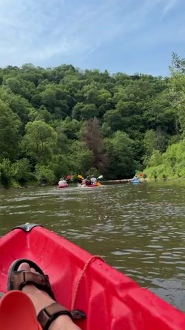 From the perspective of someone sitting in a red kayak on a river, a group of people can be seen further ahead on the river in kayaks and inflatable tubes. The group seems to be clustered around some debris in the river. The river is surrounded by a dense, green forest on both sides, under a blue sky with wispy clouds.