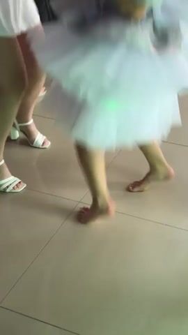 A person's bare feet are visible as they dance in a white tutu. Another person's feet in white sandals are visible on the left. The floor is tiled. The scene is lit with purple and white lights.