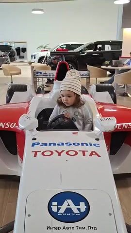 A young child wearing a striped beanie sits in a red and white Formula 1 race car replica. The car has "Panasonic TOYOTA" and "DENSO" logos on it, as well as a blue and white "IAI" logo at the front. The child is holding the steering wheel and appears to be looking forward. The background shows a car dealership with other vehicles and furniture visible. The child's mouth is open as if speaking or reacting to something. The child's hands are on the steering wheel.