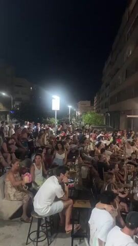 The video shows a crowded outdoor scene at night, possibly a street or plaza lined with buildings. Numerous people are gathered, many seated at tables and chairs, suggesting a social gathering or an outdoor dining area. There are streetlights illuminating the area, and some buildings have illuminated windows. People are dressed in casual summer clothing, and the overall atmosphere appears lively and active. In the background, a sign above a building appears to be for a business, possibly a bar or restaurant. The camera pans slightly, showing the density of the crowd and the general setting.