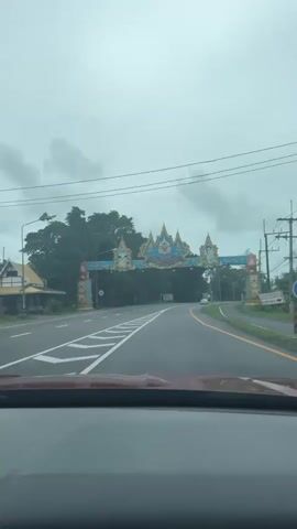 The video shows a view from inside a vehicle driving down a road. Ahead, there is a large, ornate archway spanning the road. The archway is decorated with blue, gold, and red elements, and features what appear to be images of royalty or important figures. The road has multiple lanes and white lane markings. On either side of the road are trees and vegetation. The sky is overcast and appears cloudy. There are utility poles and wires visible along the side of the road. The vehicle is moving forward, and the archway is approaching.