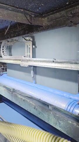 The video shows an industrial setting, possibly a printing or coating factory, where a large blue roll of material is being processed by a machine. The roll is moving forward on a system of rollers, with some machinery positioned above it. The scene is dimly lit, highlighting the metallic and aged look of the equipment. There are no people visible in the shot.