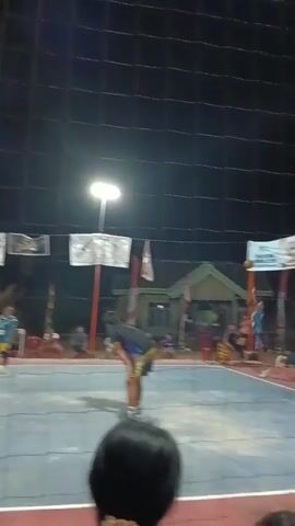 A volleyball game is in progress at night under artificial lighting. The court is visible with a net dividing it. Several players are on the court, some in action and others observing. Spectators are visible behind a net in the foreground. Banners are hung in the background. The overall atmosphere suggests a local sporting event.