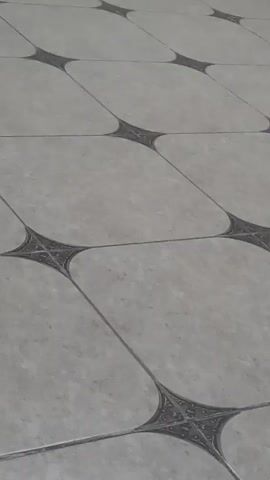 A close-up, low-angle shot captures a tiled floor. The tiles are square with a light grey, speckled pattern. Darker grey, intricate star-like designs are present where the corners of four tiles meet. The camera is angled upwards, moving slowly across the floor. The lighting is even and appears to be artificial. There are no people or other objects visible in the frame.