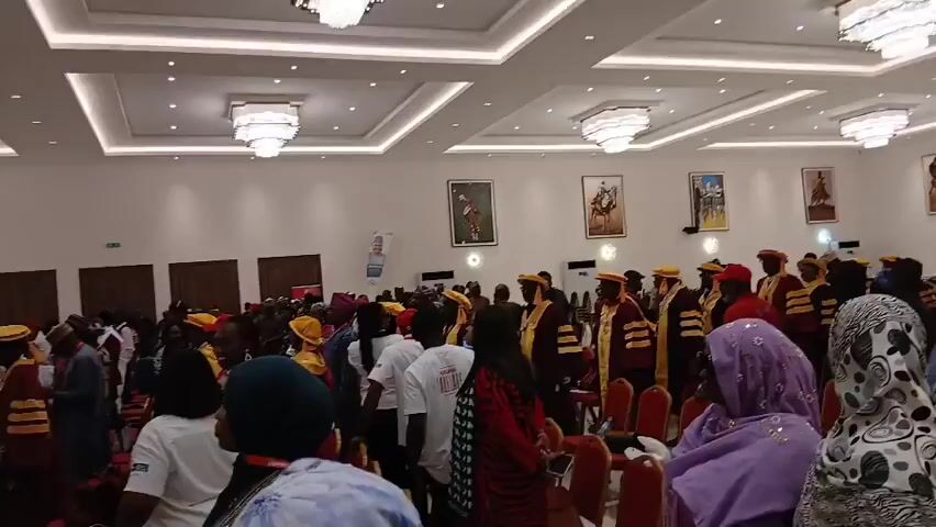 The camera films a large, well-lit room filled with people. Most are standing, wearing a variety of clothes. Many people are wearing yellow and red academic robes and caps, suggesting a graduation or similar ceremony. The room is adorned with several large paintings depicting people on horseback and other scenes. The ceiling has recessed lighting and large, square chandeliers.  The scene shows a bustling, celebratory atmosphere.