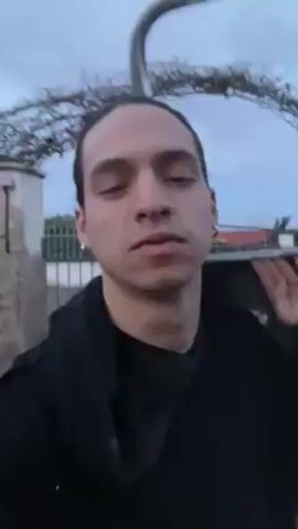 Here's a description of the video:

A person with dark hair pulled back into a bun, wearing a black jacket and small hoop earrings, is holding the camera and filming themself outdoors. The person makes a disgusted facial expression, wrinkling their nose and showing their teeth.  Behind them is a gate or archway structure with some greenery, a wall, and a vehicle covered with a tarp in the background. The sky is visible and appears to be early evening or dusk.