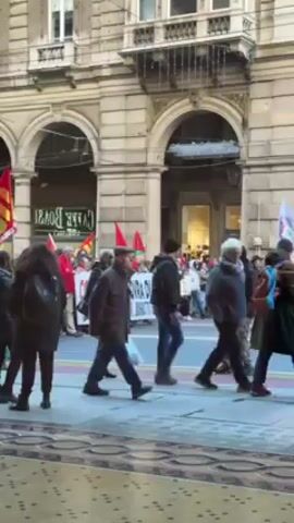 A crowd of people is walking along a sidewalk next to a street. They are carrying flags and banners, some of which are red and yellow. The buildings in the background have arched windows and decorative stonework. The crowd is moving in one direction, and some people are looking around and talking to each other.