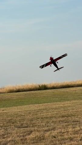The video shows a remote-controlled airplane in flight against a clear sky with scattered clouds. The airplane is red and black with white accents and has its landing gear deployed. It appears to be performing a maneuver, possibly a climb or a stunt, as it is tilted at an angle. The background consists of a grassy field with dry, tall grass in the foreground and a distant landscape with rolling hills and what appear to be wind turbines. The scene is set outdoors during what seems to be the daytime, with soft lighting suggesting it might be late afternoon or early morning. There is no visible text within the video. The overall impression is one of outdoor recreation and hobbyist activity.