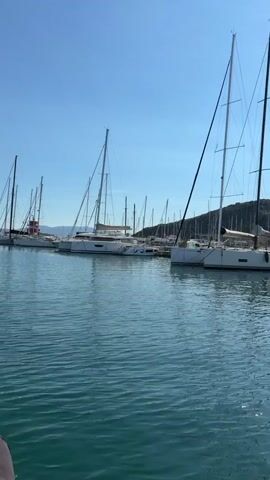 The video shows a marina with several sailboats docked. The water is calm and blue, and the sky is clear. The sailboats are white with tall masts. In the background, there is a hill covered with trees and buildings. The camera pans slowly from left to right, showing more of the marina. There are no people visible in the video.