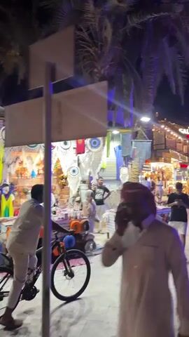 The camera follows the activity on a tourist shopping street at night, somewhere with a Middle-Eastern feel. Palm trees line the street, lit up with purple-blue light. People stroll down the pedestrian path, some looking at their phones, others in groups, some on bicycles. There are several small shops selling trinkets and souvenirs, and signs above the storefronts such as “Sea Cave” and “Salt&Sky Hotel”. The street is crowded, with people moving in both directions. The overall impression is of a vibrant and bustling tourist destination in the evening.