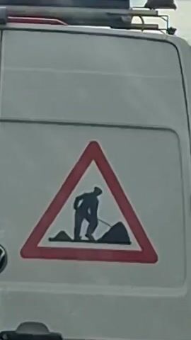 A close-up shot shows the rear of a white van. A triangular warning sign with a red border depicts a silhouette of a person digging with a shovel. To the left of this sign, a blue circular sign with a white arrow pointing down and to the left is visible. Below these signs, a rectangular red sticker with white text reads "How's my Driving?". The van has red and white striped reflective tape along the bottom. A yellow warning light is mounted on a rack above the van. The VW logo is visible on the rear door.