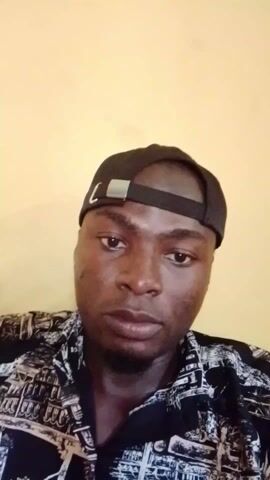A young man with a dark complexion is featured in a close-up, chest-up shot. He is wearing a dark baseball cap backward, and a black and white patterned short-sleeved shirt. His short, dark hair is visible beneath the cap. He has dark eyes and a generally neutral to slightly contemplative expression, looking directly into the camera. Throughout the short clip, he makes subtle, slight head movements and blinks occasionally. The background is a plain, light yellow or beige wall, indicating an indoor setting. The lighting is even and consistent, suggesting either daytime indoors or a well-lit room. There are no other specific objects or discernible text visible in the frame. The man appears to be directly addressing or acknowledging the camera, possibly recording a personal video or selfie.
