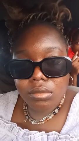 A person with brown skin and dark hair is wearing black sunglasses, a light-colored ruffled shirt, and a necklace. They are in a car with a driver and another person in the background. The person is looking directly at the camera. The background is slightly blurred.