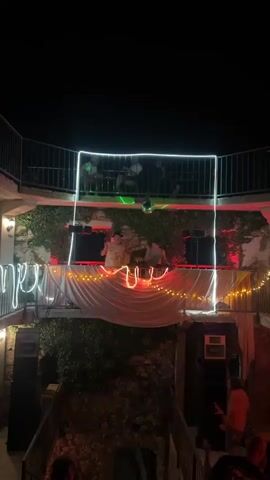 This is a vertical, nighttime shot of an outdoor party or event. In the foreground, on a lower level, a person in a pink shirt gestures with their hands while looking upwards. To the left and right of this person are large black speakers. Above this lower level is a raised platform where three people are gathered around DJ equipment. The platform is decorated with white draping and strings of orange lights. A white neon light outline frames the DJ area. Green lights flash above the DJ booth. Further up, on a balcony or upper level, several silhouetted figures are seated at tables. A disco ball hangs in the center, reflecting light. The background consists of dark foliage and stone textures.