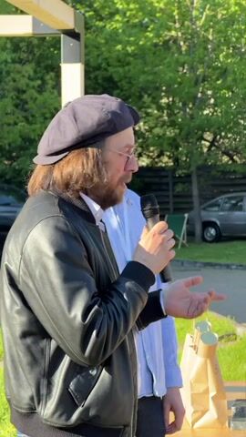 The video shows a man in a black leather jacket and a dark-colored cap speaking into a microphone in an outdoor setting. He gestures with his hands as he talks, and another man in a light blue shirt stands behind him. A table with a brown paper bag and other items is in front of them. The camera then pans out to show a group of people standing on a brick pathway in a grassy area with trees and parked cars in the background. The sky is clear and blue.