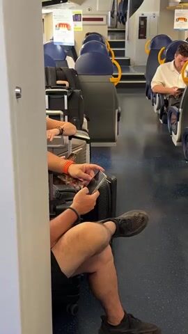 The video shows the interior of a train carriage with passengers. In the foreground, a person wearing black shorts and dark sneakers is seated, holding and looking at a smartphone. They have an orange wristband on their left wrist and a blue bracelet on their right wrist. Luggage, including a black suitcase and a grey carry-on bag, is visible next to them. Another person's arm, wearing a watch, is visible slightly behind them. In the background, other passengers are seated, and luggage is stored. The seats are blue with yellow grab handles. The train carriage appears to be in motion.