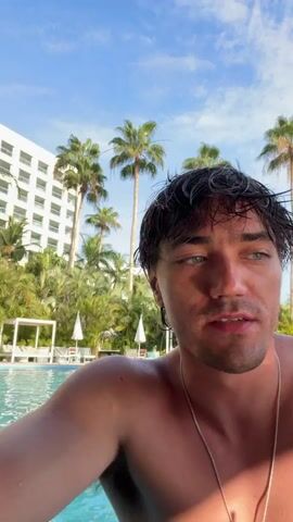 The video shows two men at a pool bar in a tropical resort. The first man, with wet dark hair and striking blue eyes, stands in the water with a large resort hotel and palm trees in the background. He is shirtless and looks at the camera. Then the video shifts to a second man with a beard who is also shirtless in the pool, holding up a pair of sunglasses. The video cuts back to the first man, who is now holding a green cocktail garnished with a slice of lime. He smiles at the camera.