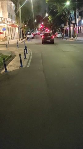The video shows a first-person perspective from someone moving along a street at night, likely on a bicycle, starting with a close-up of a bicycle handlebar and then panning to reveal the surroundings.

The scene depicts an urban or suburban street at nighttime. On the left side of the street, there is a building with outdoor seating areas featuring red umbrellas and tables, suggesting a restaurant or cafe. Blue, short, cylindrical bollards separate the pedestrian area from the roadway.

Further down the street, red traffic lights are visible, and several cars are present, some parked and others seemingly in traffic. The road itself is asphalt, marked with double white lines. The sky is dark, indicating night, and various artificial lights illuminate the area.

A notable detail is the visible text "La Veneciana" on the building on the left, which is likely the name of the establishment. The activity involves continuous movement along the street. The weather appears clear, with no rain or fog.