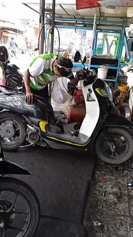 The video shows a mechanic in a green and white soccer jersey working on a black and white Honda scooter in a garage. The scooter is raised on a lift and partially disassembled. Tools and parts are scattered around the scooter and the garage floor. The mechanic is leaning over the scooter, then straightens up to look at it. Other motorcycles are visible in the background. The garage appears to be open to the outside, with street traffic visible in the distance. The floor of the garage is made of black rubber mats.