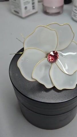 The video shows a close-up shot of a white flower decoration with a pink gem in the center, placed on top of a black cylindrical object. The flower appears to be made of translucent material with gold outlining. The background is blurred but shows some containers and objects, suggesting an indoor setting, possibly a crafting or display area. The lighting is soft, highlighting the details of the flower and gem. The camera slowly pans around the object, offering different angles.