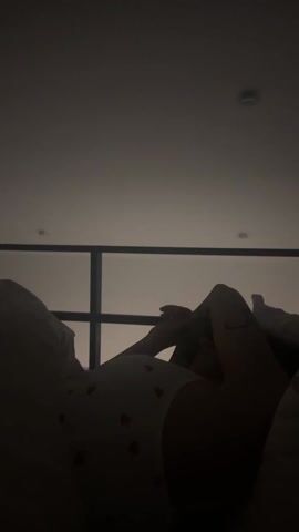 A woman lies in bed in a dimly lit room, with her legs raised and hands clasped together in her lap. She is wearing what appears to be a white top with red heart patterns.  A window with a distinct cross-shaped mullion is behind her, letting in some light, and the ceiling is visible above. The lighting is overall quite low, emphasizing silhouettes.