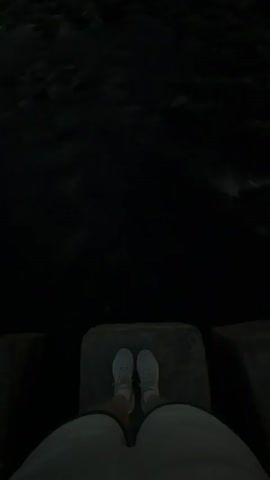 The video shows a person's legs and feet standing on a concrete structure. The person is wearing white sneakers and light-colored shorts. They are standing with their feet slightly apart, facing forward. The area below the concrete structure is dark, appearing to be water. The lighting suggests it is nighttime or very early morning, with the dark water reflecting some light. The concrete structure appears to be part of a larger barrier or walkway. The overall scene is dark and somewhat ominous, with the focus on the person's feet and the dark expanse of water before them.