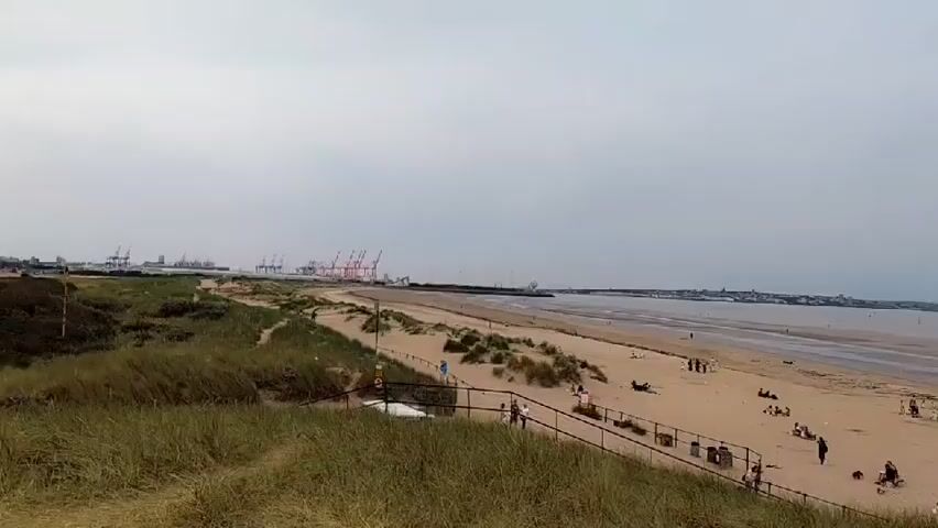 This video captures a wide shot of a sandy beach on a cloudy day. In the foreground, tall, dry grass grows on a dune. A wooden fence separates the dune from the beach. Several people are scattered across the sand, some sitting or lying down, while others walk along the shore. Beach umbrellas and a yellow windbreak are visible. In the distance, the sea stretches out, with a low tide revealing exposed sandbanks. Several figures can be seen walking on the sandbanks in the water. To the far left of the frame, large, red industrial cranes are visible, suggesting a nearby port or industrial area. A few buildings are faintly visible along the coastline in the distance. A dark, slender structure, possibly a lighthouse or marker, stands in the sea. The overall atmosphere is relaxed and casual, with a few individuals enjoying a day at the beach.