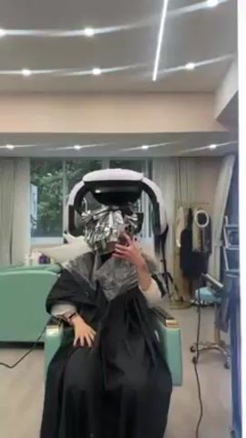 A person sits in a salon chair, covered with a black cape and foil in their hair. A white hair-processing machine is positioned over their head. The person is holding a phone and filming themselves, looking at the screen and holding up two fingers. The salon has a light pink ceiling with round and linear light fixtures, a light-colored sofa, a large window with a view of trees, and a gold clothing rack with various garments. A vanity with a mirror and equipment sits to the right. The camera view appears to be from a large mirror in front of the person.