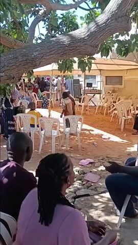 Here's a description of the video:

The scene takes place outdoors under the shade of a large tree. The focal point is a gathering of people, most seated on white plastic chairs under a canopy. Some people are standing around the periphery. A small stage area is set up, with a keyboard and speakers visible. Two children are dancing and moving around in the open space between the seated crowd and the stage.  

In the foreground, three people are seated on a low structure, facing the gathering. The person in the middle has dark hair styled in long braids. Discarded styrofoam food containers are scattered on the ground.

The overall atmosphere appears lively and celebratory. The scene is captured with a slightly shaky hand-held camera, providing a ground-level perspective.