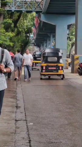 The video shows a street scene with several people walking on a wet pavement next to a road. In the foreground, a person wearing a grey patterned shirt and dark pants walks towards the camera, carrying a backpack and looking at their phone. Further down the street, a man in a white t-shirt and jeans walks away from the camera. Behind him, two auto-rickshaws are visible on the road, one black and yellow and the other yellow and black. The background features trees, a pedestrian overpass, and structural elements of what appears to be an elevated railway or highway. The overall environment suggests an urban setting.