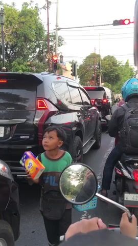 The video captures a street scene in Pagerwojo, East Java, likely during the late afternoon or early evening. The lighting suggests it's neither midday nor nighttime, and the sky appears overcast or hazy, indicating potentially moderate weather conditions. The setting is an urban street with vehicular traffic, characterized by a traffic light visible in the background, suggesting an intersection.

The primary subjects are a young boy and several vehicles. The boy, dressed in a green and brown t-shirt, stands on the side of the road, holding a box of tissues. He appears to be a vendor, possibly selling the tissues to drivers stuck in traffic. His expression is neutral or slightly curious, and his posture is alert as he looks towards the camera.

In the foreground, a motorcycle mirror is visible, implying the footage is captured from a motorcycle. A person on the motorcycle, wearing a blue helmet and a backpack, is also present. Several cars are stopped in traffic, including a black SUV in the middle ground. The vehicles suggest a typical urban commuting environment.

The activity taking place is the observation of street vending within a traffic jam. The boy is engaged in his work, and the overall scene is one of everyday life in a busy Indonesian street.

Notable details include the presence of traffic lights, the style of the vehicles, and the boy's apparent role as a vendor. The general atmosphere seems to be one of routine activity rather than a specific event.

Visible text includes branding on the tissue box the boy is holding. The box is orange and blue and has "Nic" written on it. The license plate of the black SUV has "BE" visible on it. The license plate of the motorcycle is partially visible and appears to have "W 420" on it.