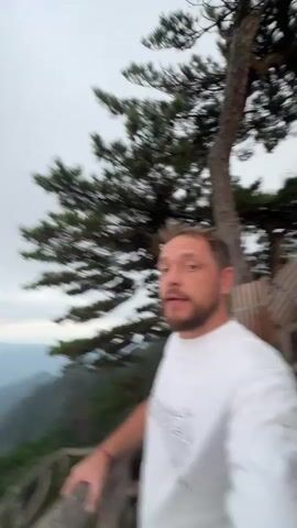 A man in a white t-shirt stands on a wooden walkway overlooking a vast mountain range. The mountains are jagged and covered in lush green trees. The sky is overcast and a light pink hue can be seen in the distance. The man turns his head to look out at the scenery, then back towards the camera. He gestures with his hands as if he is speaking. The camera pans to show the full grandeur of the mountains.