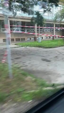 The video shows a view from inside a vehicle looking out a window. Outside, there is a building with multiple windows and a surrounding area with trees, grass, and pavement. There are some green and white striped bollards and a red and white striped barrier visible. The video captures movement, suggesting the vehicle is in motion. The building appears to be an industrial or institutional structure. Text on a sign in the distance reads "PRADELNA CISTIRNA". A fly is visible on the inside of the window.