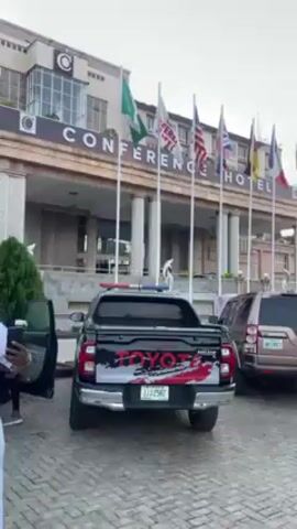The video shows the exterior of Conference Hotel, a multi-story building with a beige facade and many windows. The hotel's name is prominently displayed above the entrance, and several flags from different countries are flying in front. There are two vehicles parked in front of the hotel: a black Toyota pickup truck with red and white graphics and a brown SUV. Several people are walking around the vehicles and towards the hotel entrance. The sky is overcast, and the general atmosphere is calm. A man in a white outfit and patterned hat is visible, holding a phone.