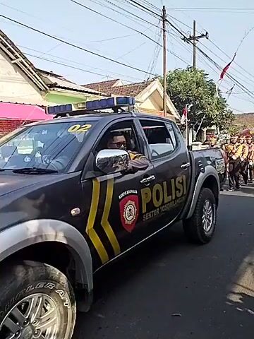 A police vehicle drives alongside a parade of young people in uniform. The police vehicle has the word POLISI printed on its side, and a police officer is visible in the driver's seat. The parade participants are marching in a single file line down a paved street. They are wearing uniforms that consist of brown shirts, dark pants, and hats. Many of them are carrying flags or banners. Spectators are visible on the side of the street, watching the parade. The scene appears to be taking place in a residential area, with houses and trees lining the street. The weather is sunny.