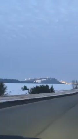 The video shows a drive on a road alongside a snow-covered field. In the distance, a hill is visible, illuminated by lights. A white van drives past in the opposite direction. The sky is overcast.